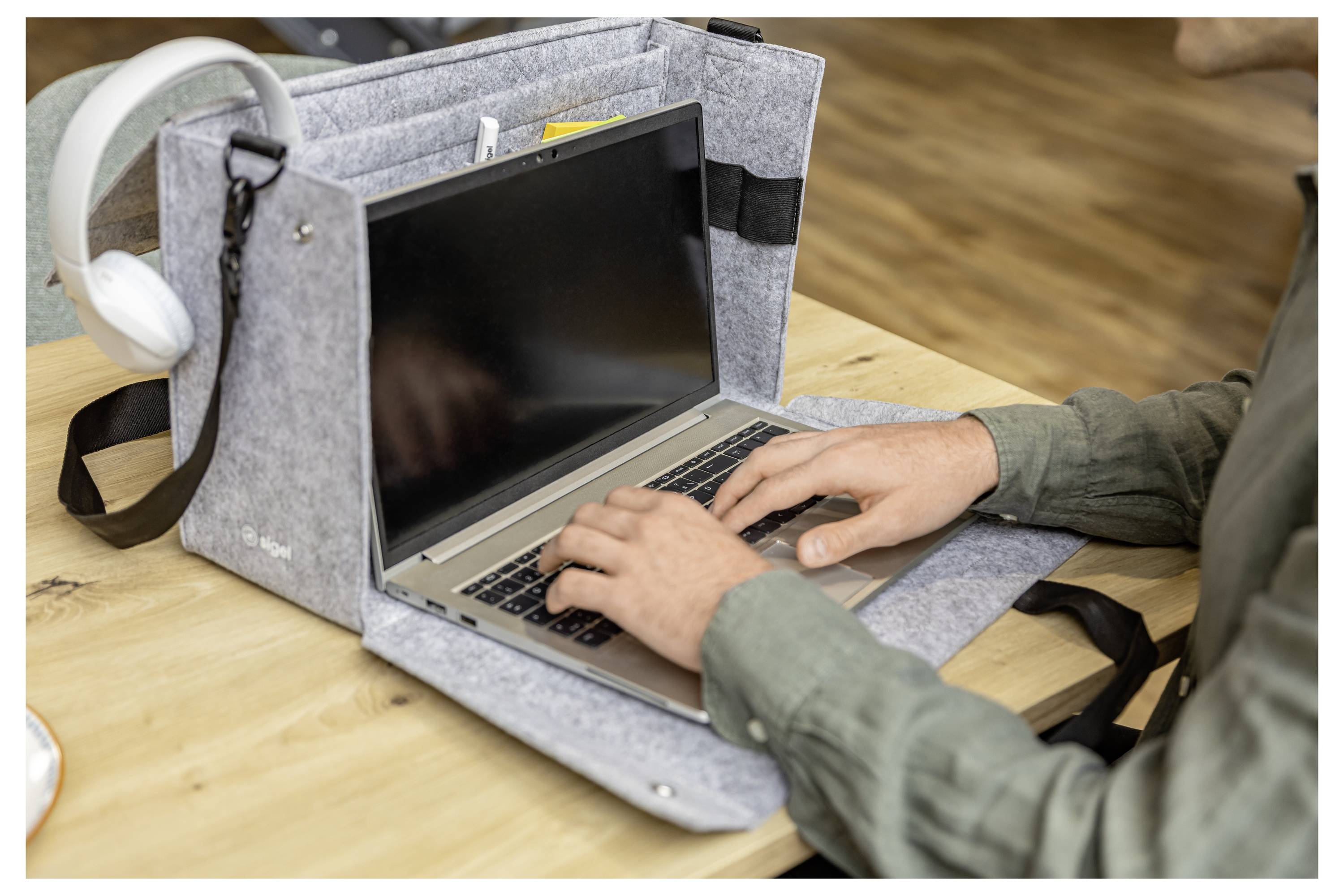 Person typing on a laptop inside a gray felt carry case on a wooden table, with white headphones hanging beside it.
