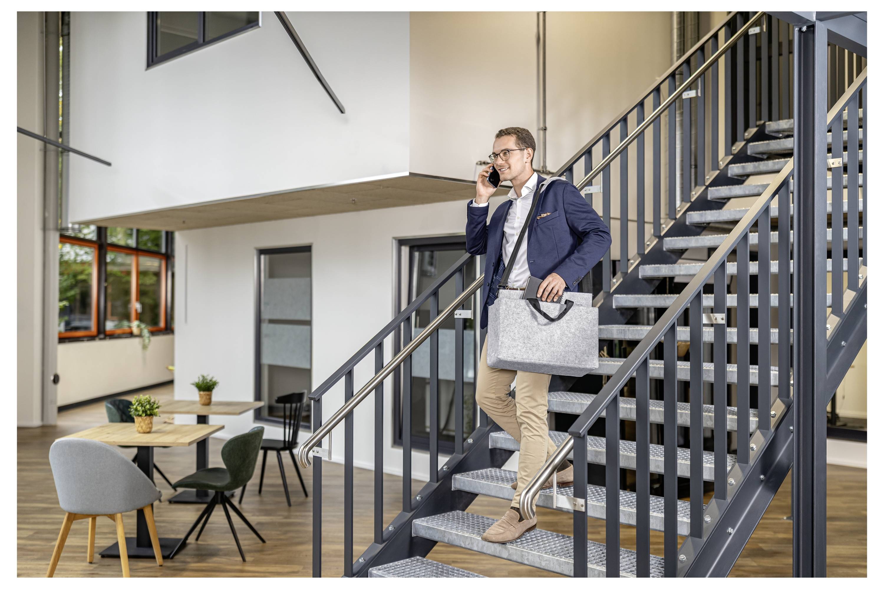 Businessman in a blazer descends an office staircase while talking on a phone and carrying a laptop bag in a modern interior.<br>