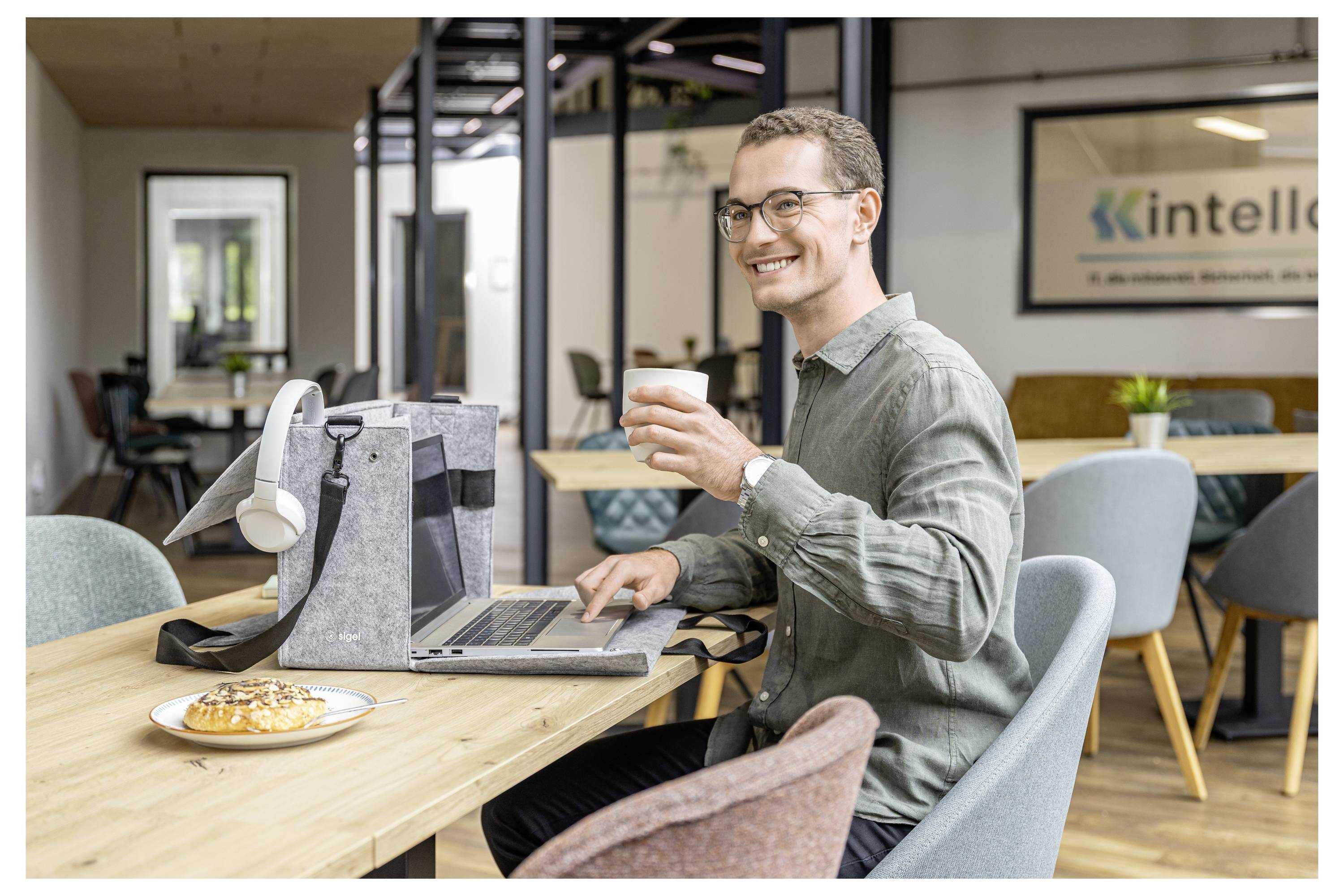 Man in a coworking space drinks coffee while working on a laptop at a shared table, with a pastry and office bags nearby.
