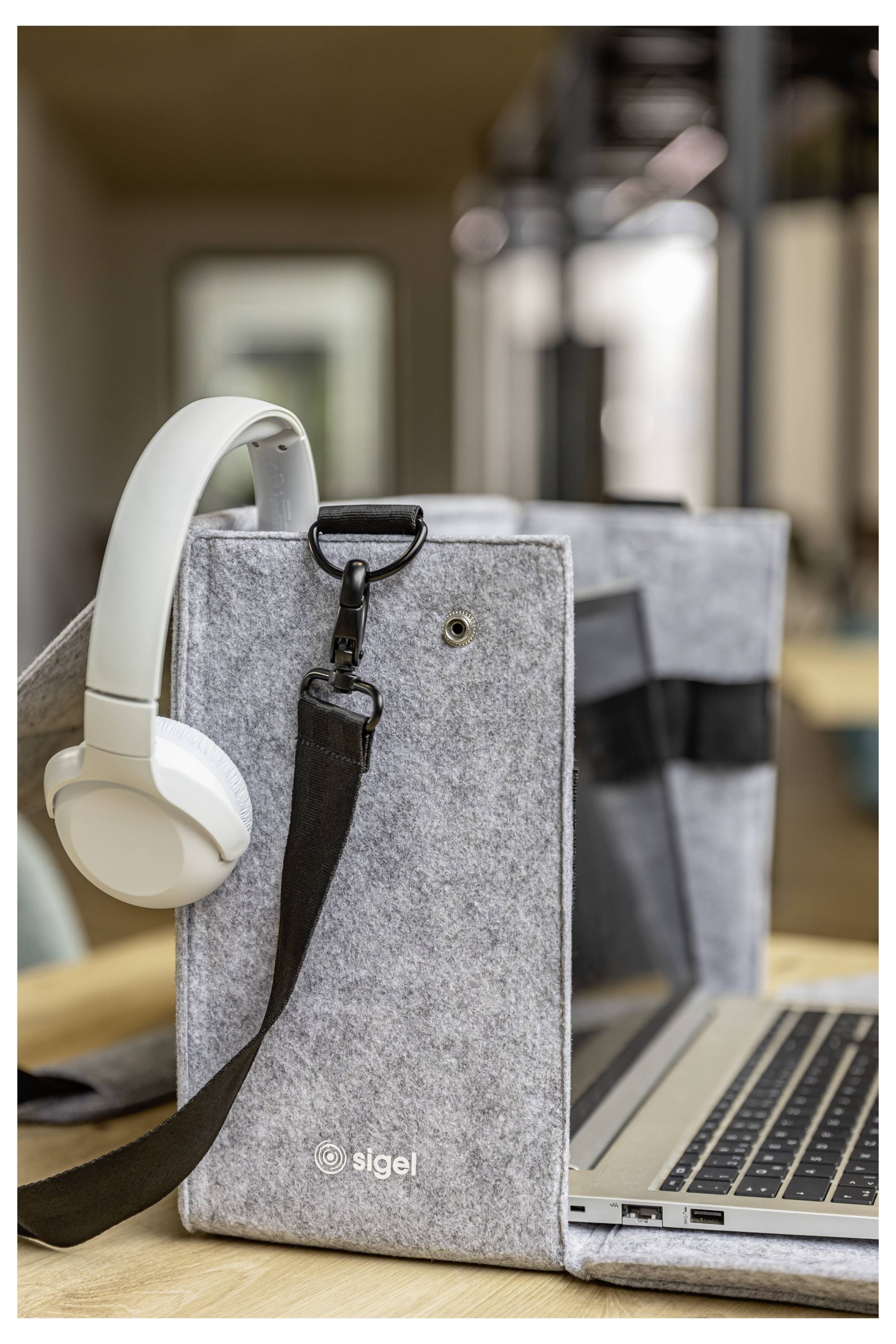 A gray felt laptop sleeve with a white headset and black strap attached, beside an open laptop on a wooden desk.