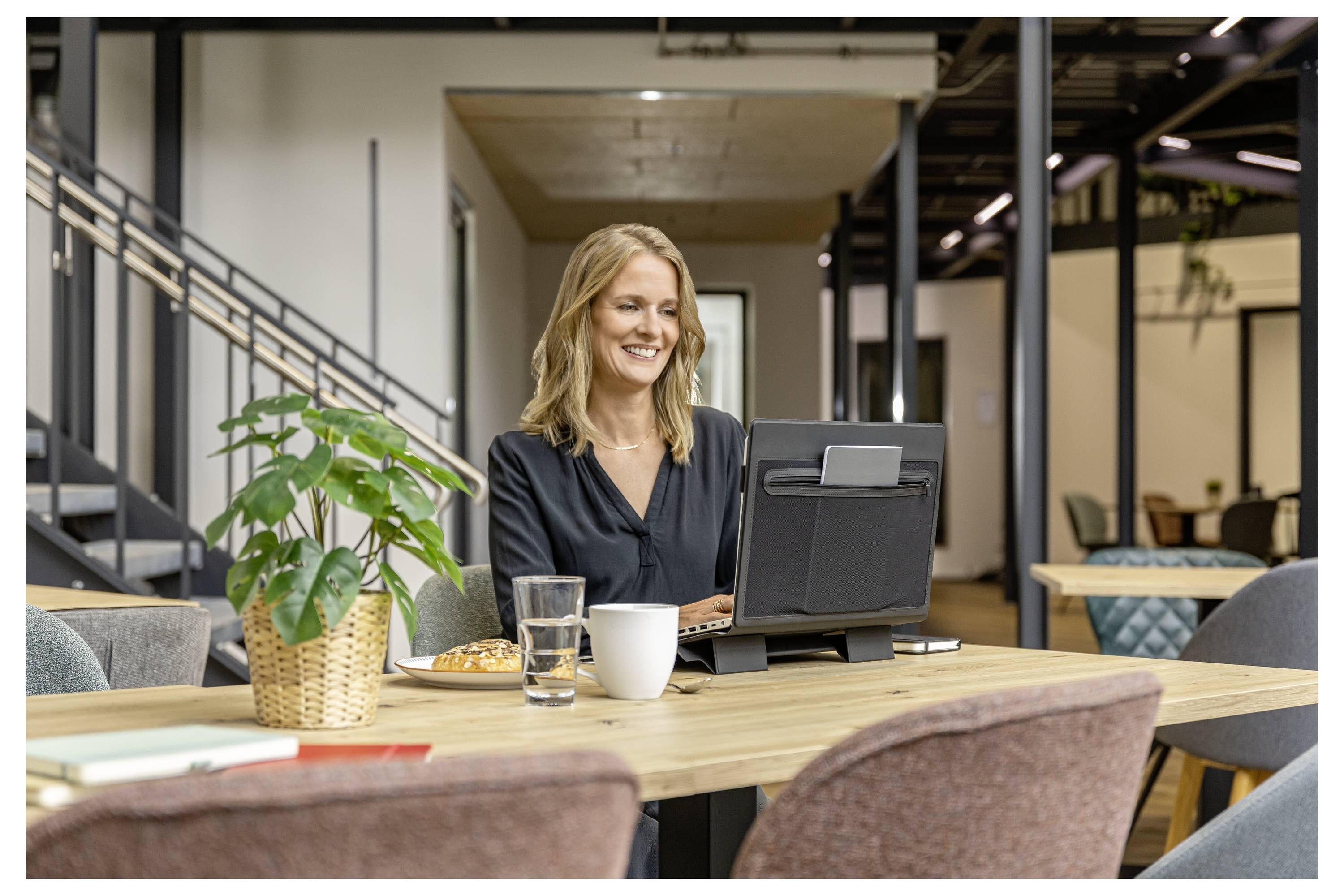Smiling woman works on a laptop at a table in a modern office lounge, with coffee, water, and a plant nearby.