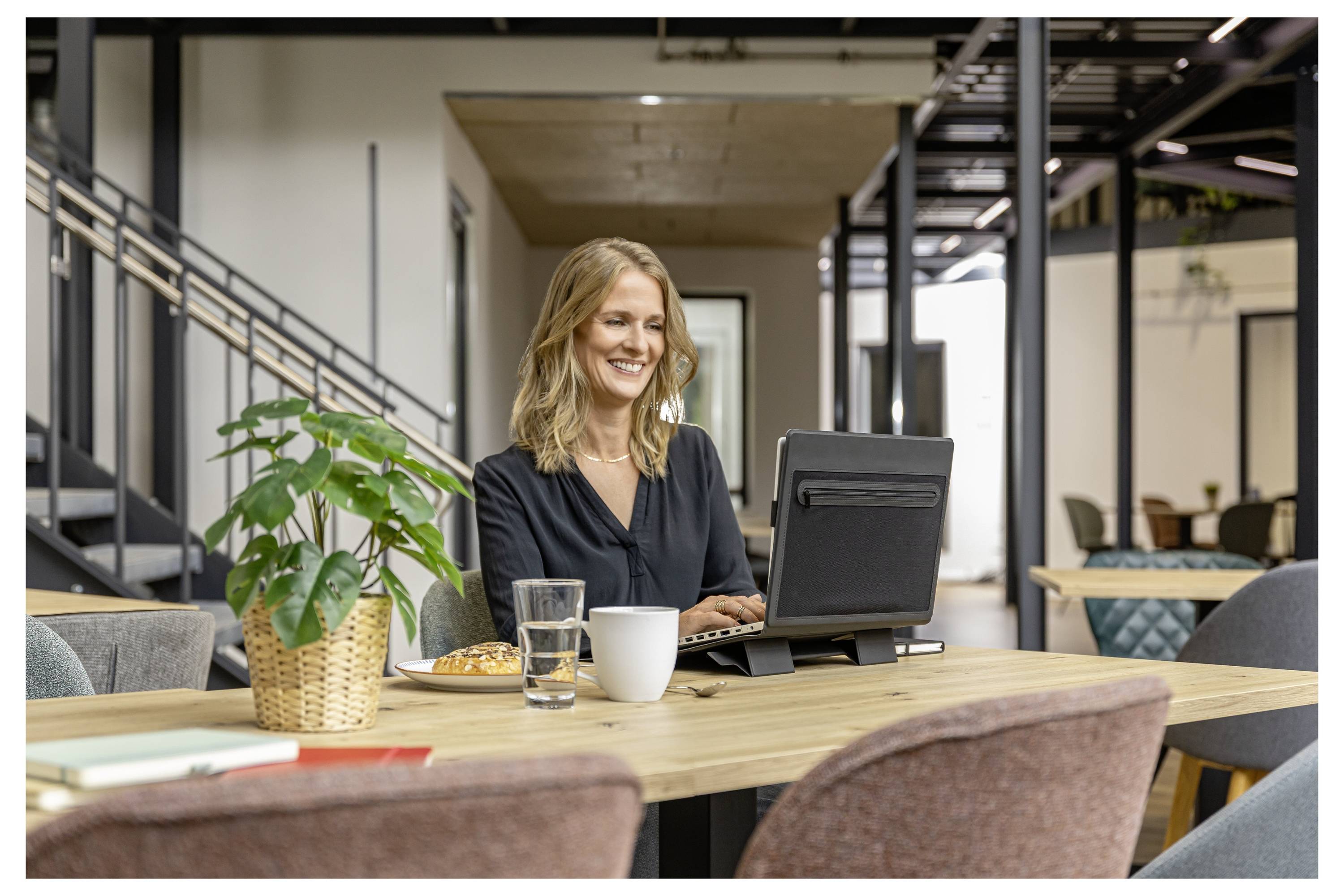 Woman smiling while working on a laptop at a table in a modern office lounge, with coffee and a glass of water nearby.