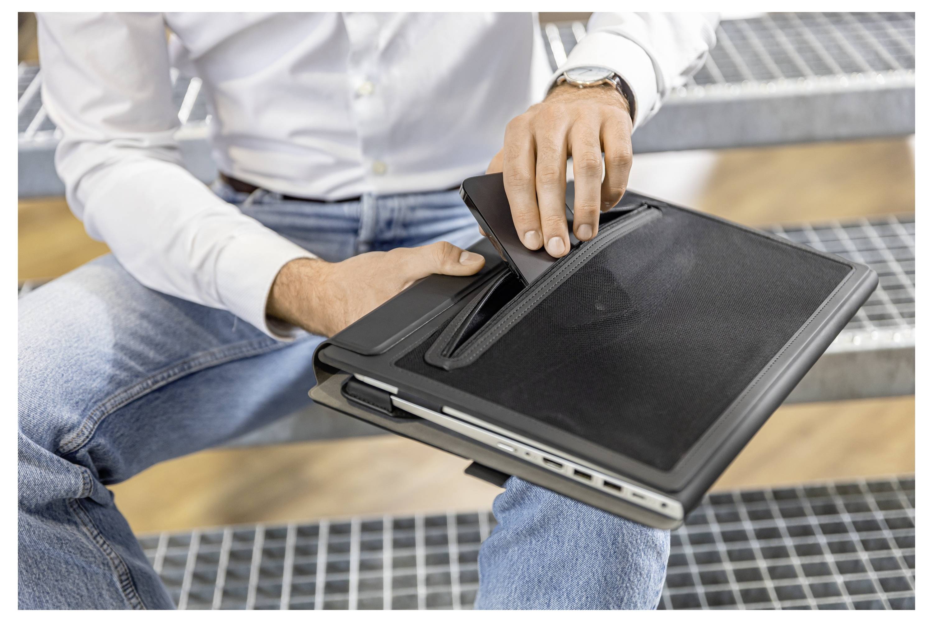 Person holding a black laptop with a zippered case resting on their lap outdoors on a metal bench.