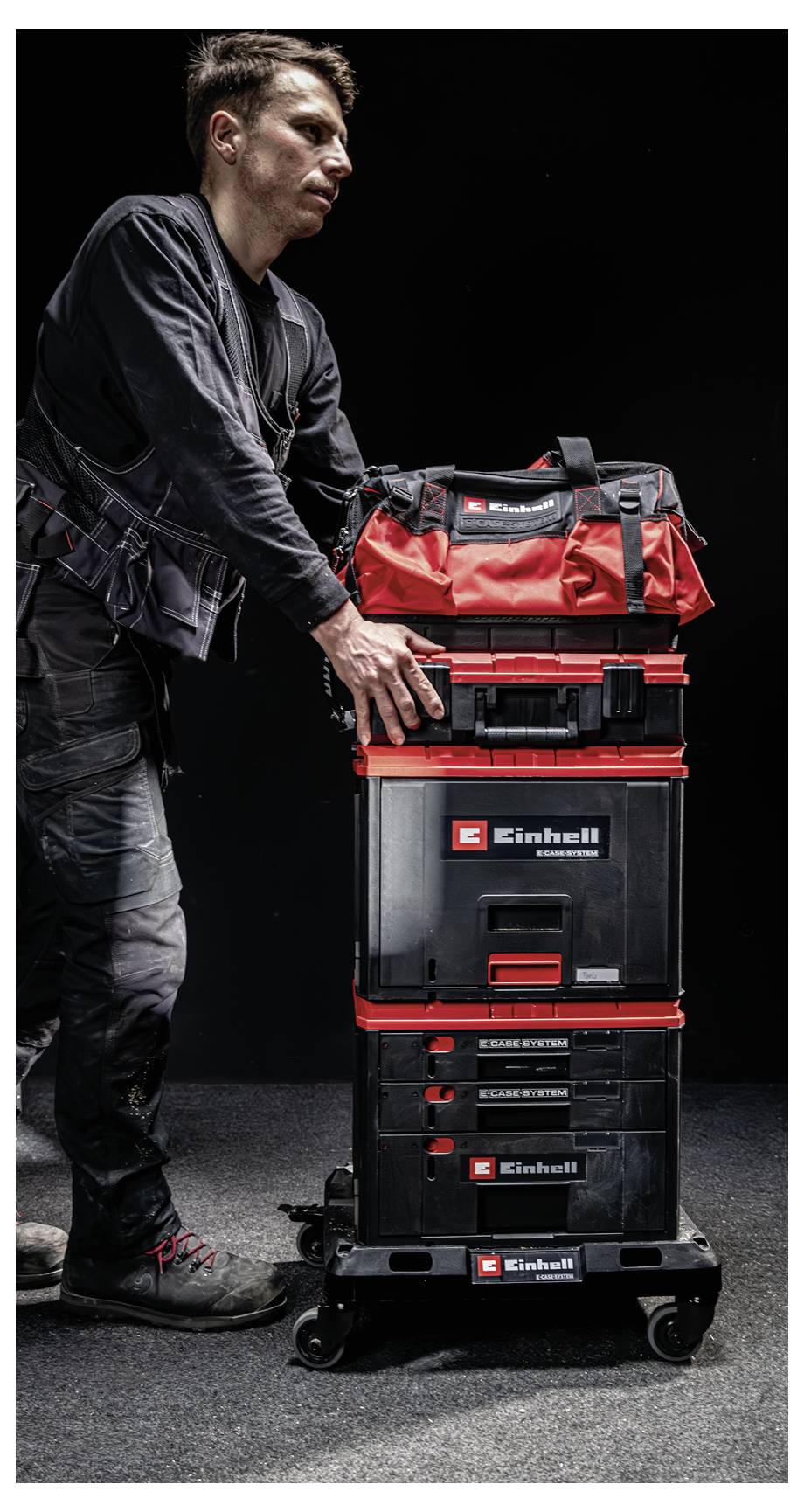 Man standing beside a stacked red and black Einhell tool storage trolley on wheels against a dark background.<br>