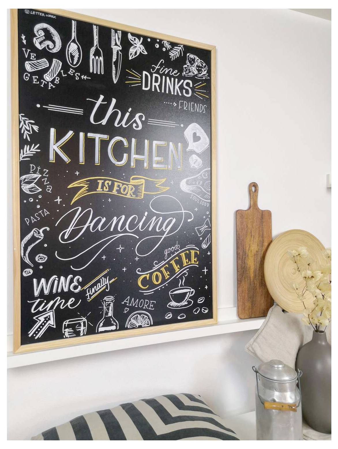 Chalkboard-style kitchen wall art reading 'this KITCHEN is for Dancing', surrounded by drink and food doodles, above a shelf with cutting boards and decor.