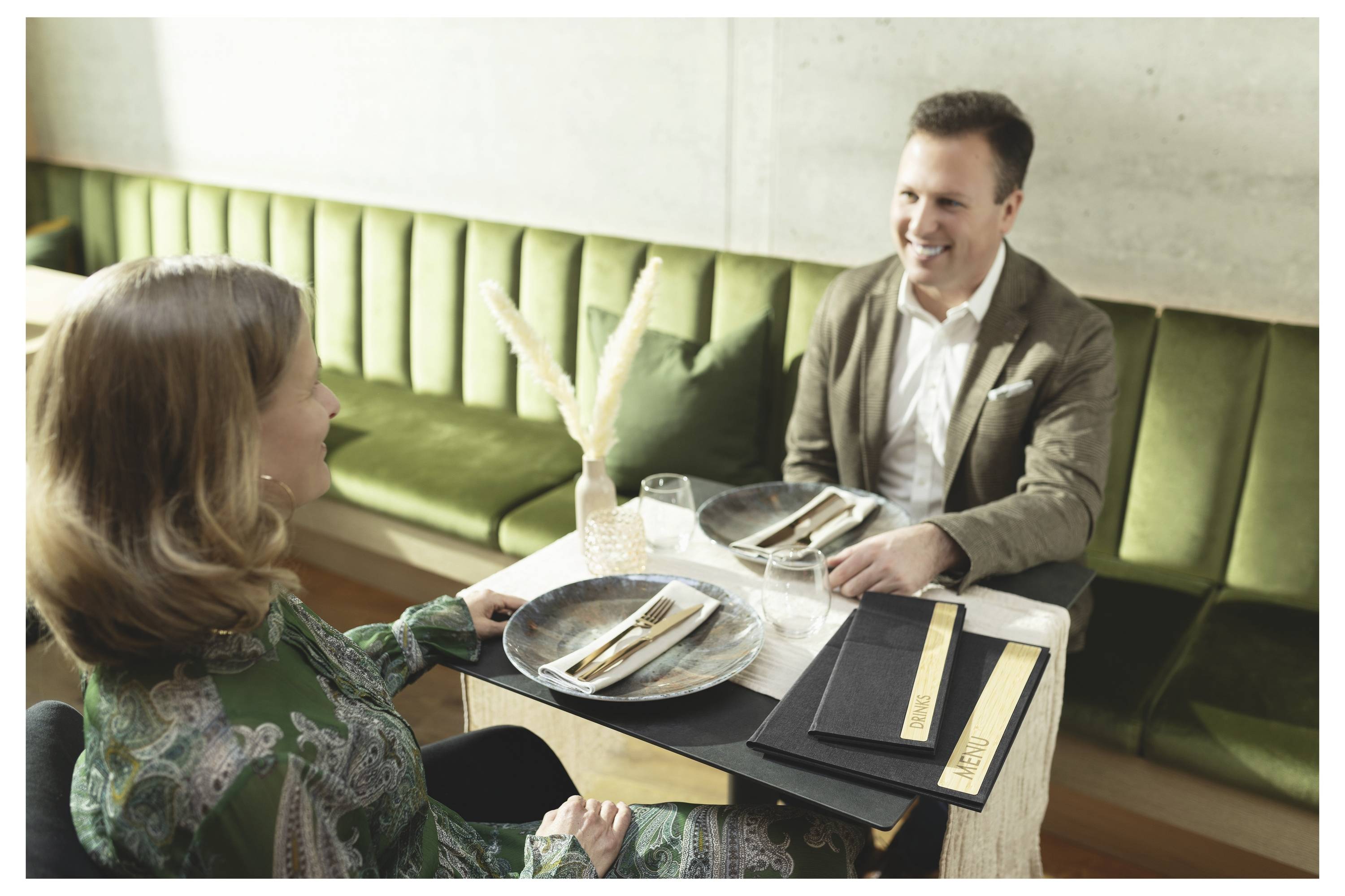 A man and woman smile while seated at a restaurant table set with plates, glasses, and menus.