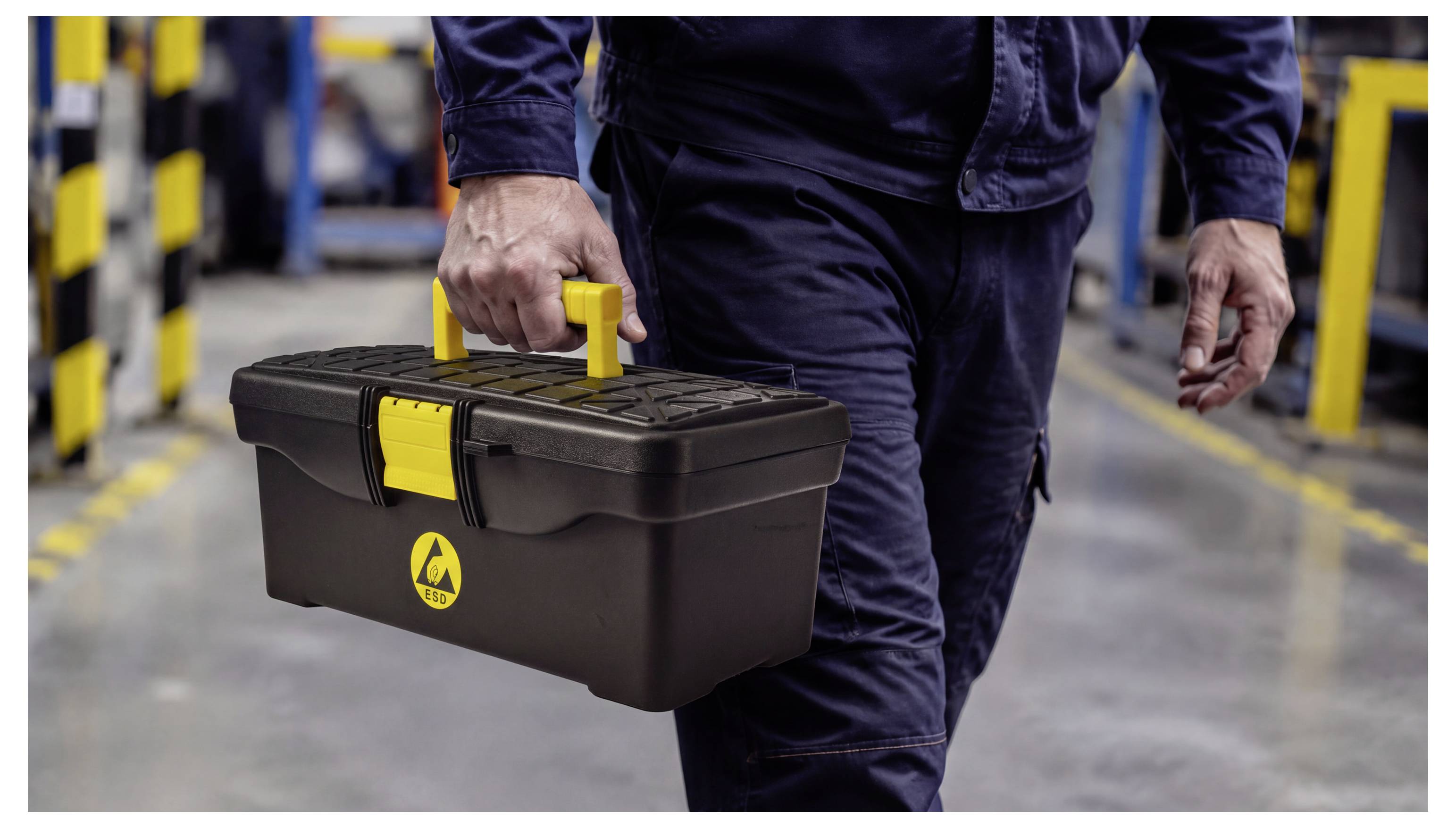 Worker carrying a black toolbox with a yellow handle in an industrial workshop aisle.