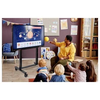 Teacher in yellow shirt points at a classroom display of the solar system while children sit and watch.