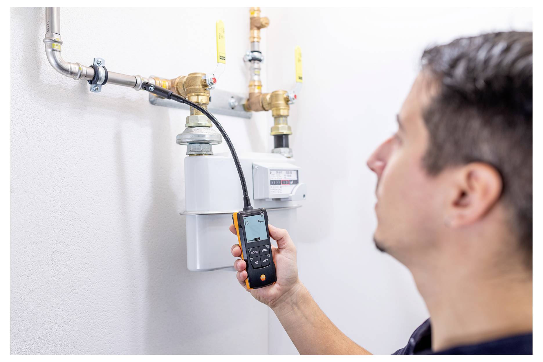 Technician holding a handheld meter to inspect a gas or utility meter with pipes and valves on a wall.