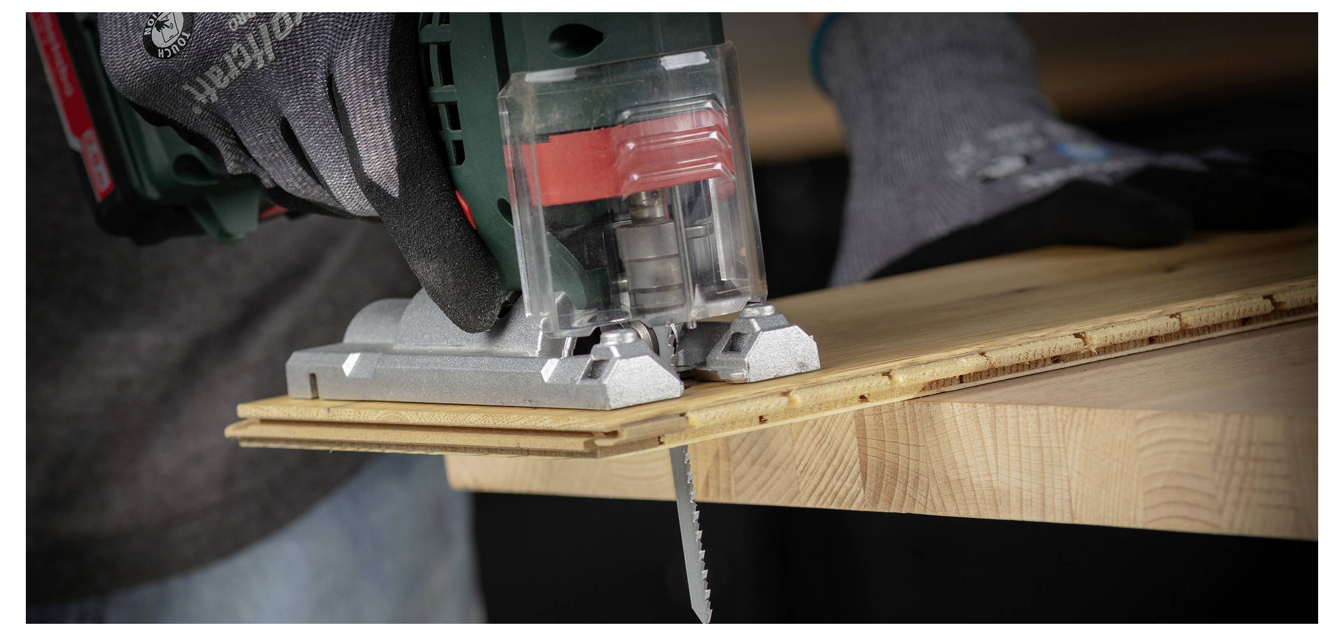Gloved hands use a jigsaw to cut a wooden board clamped on a workbench in a workshop.
