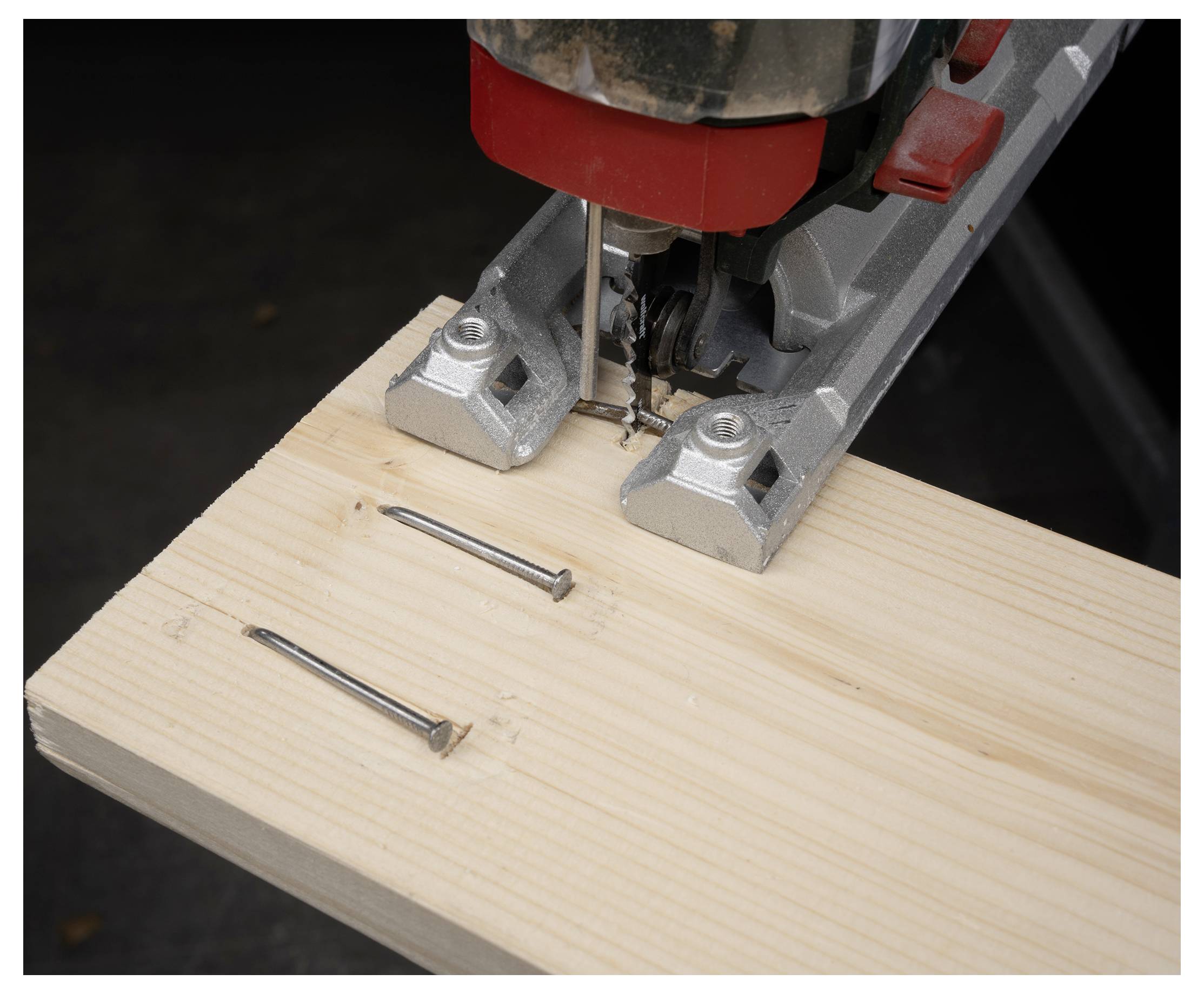 Close-up of a jigsaw cutting a light wood board with two nails nearby, showing a woodworking task in progress.<br>