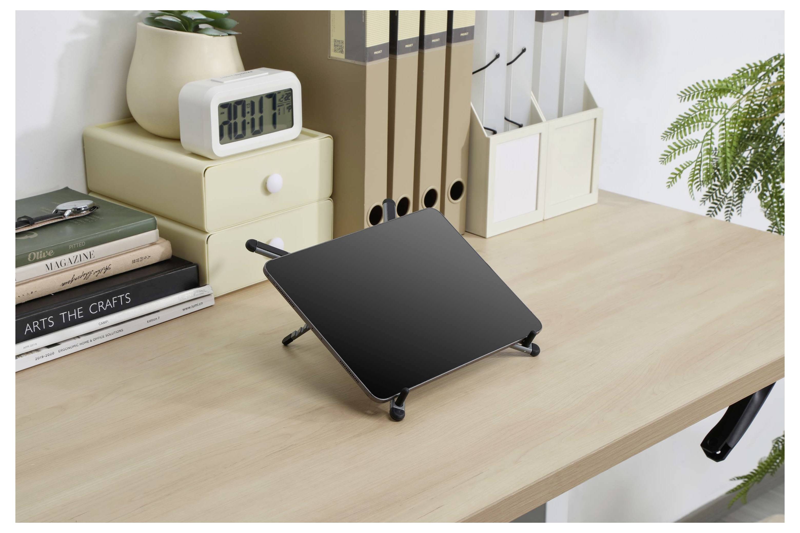 Black adjustable laptop stand on a wooden desk in a tidy home office, with books, storage boxes, a digital clock, and plants nearby.
