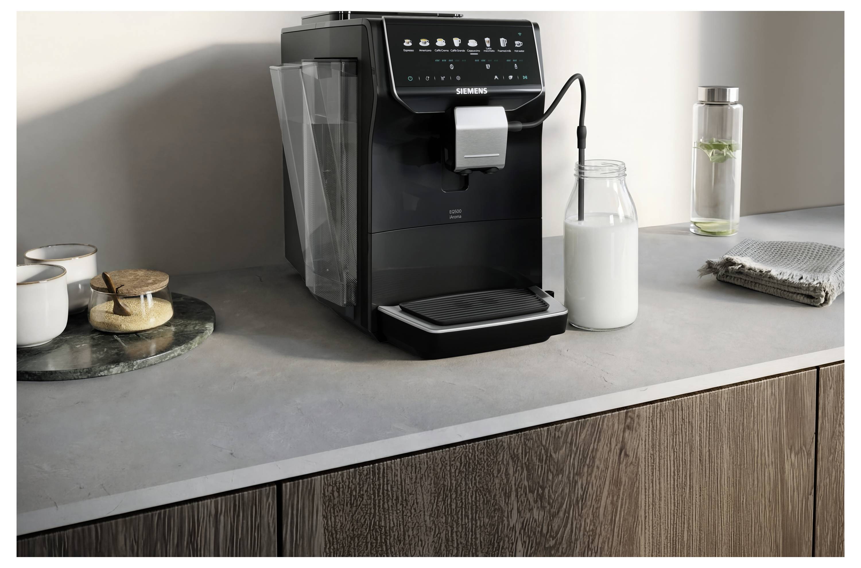 A Siemens coffee machine sits on a kitchen counter beside a milk bottle, glass water bottle, and folded cloth, suggesting a home coffee station.