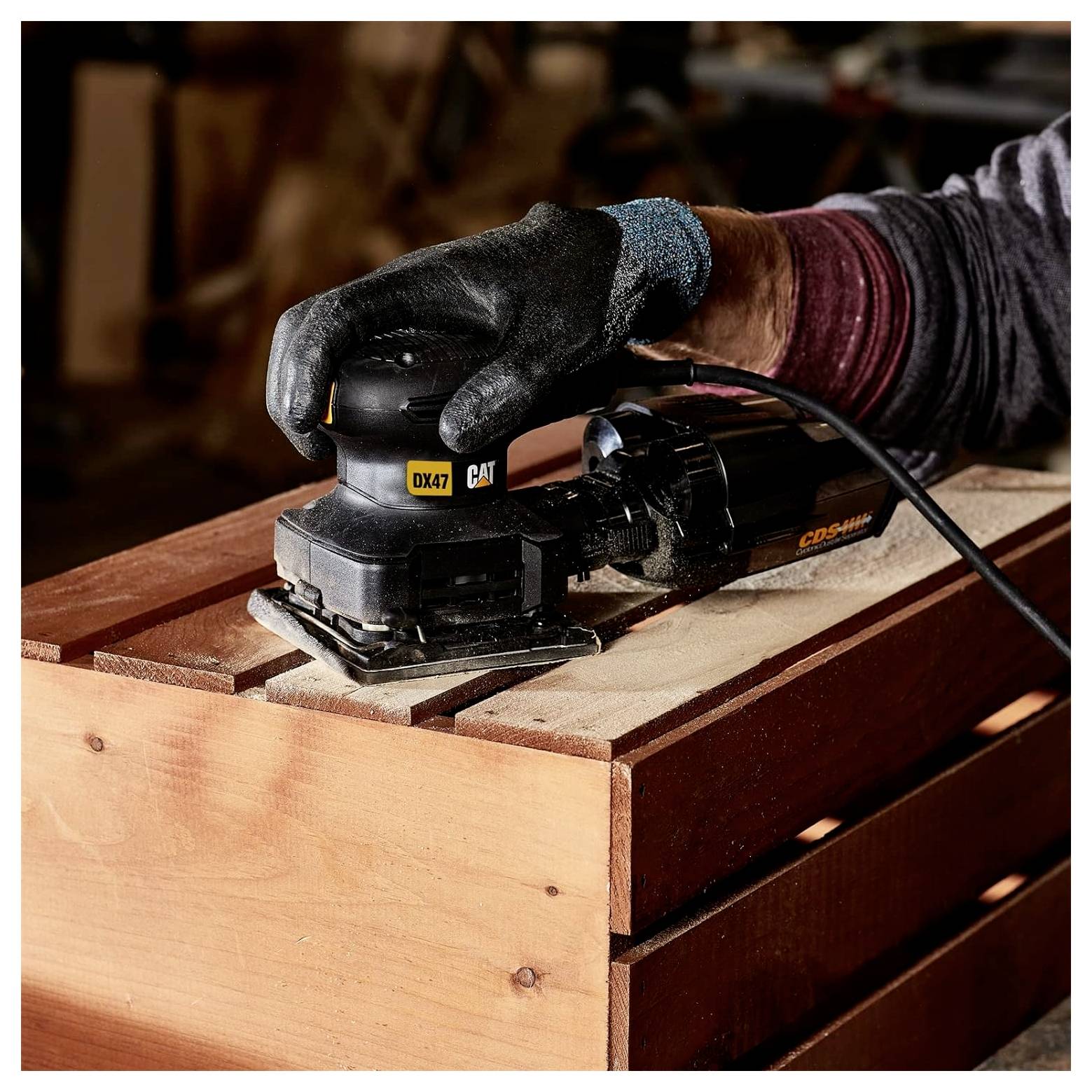 Gloved hand sanding a wooden board with a black CAT power sander on a workbench.