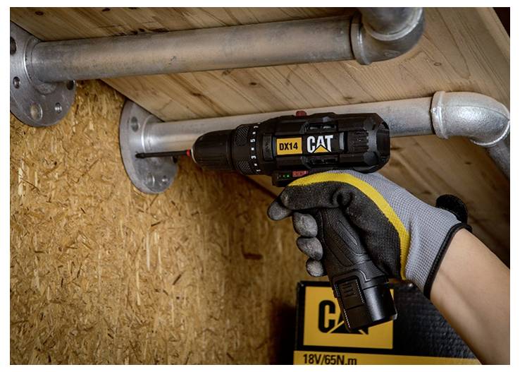Gloved hand using a CAT cordless drill to fasten a metal pipe fitting in a wooden wall framing space, with an 18V/65N·m tool label visible.