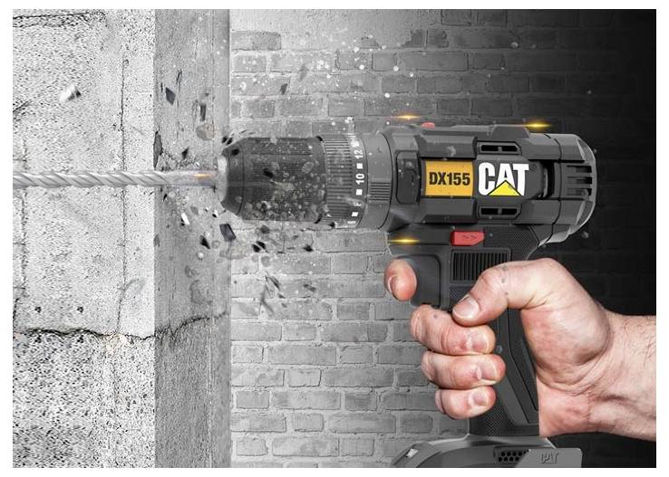 A hand operates a CAT DX155 cordless drill drilling into a concrete wall, with dust and debris flying from the bit.<br>