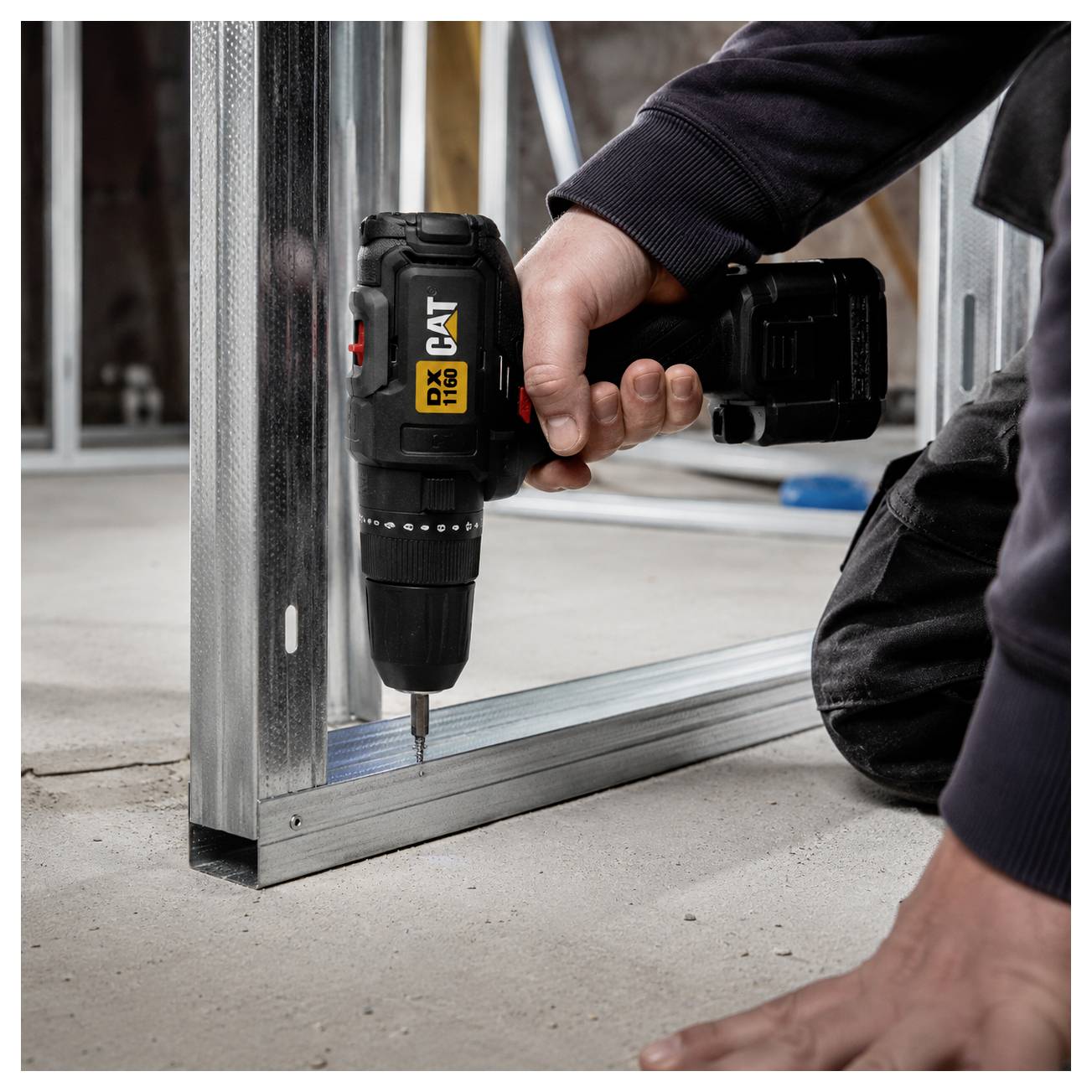 Person kneels and drills a screw into a metal frame with a cordless drill on a concrete floor.<br>