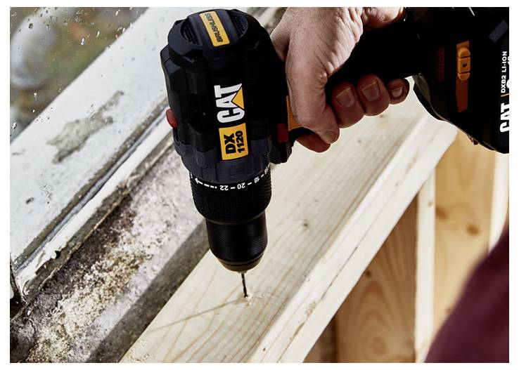 Close-up of a hand using a cordless drill to drive a screw into a wooden board beside a weathered window frame.