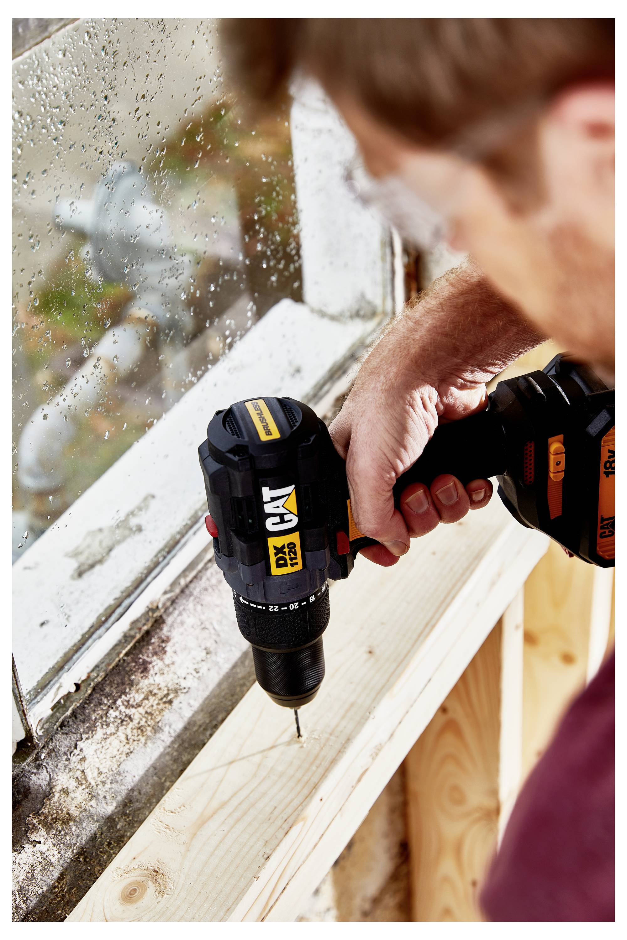 Person using a cordless drill to drive a screw into a wooden board beside a window frame.