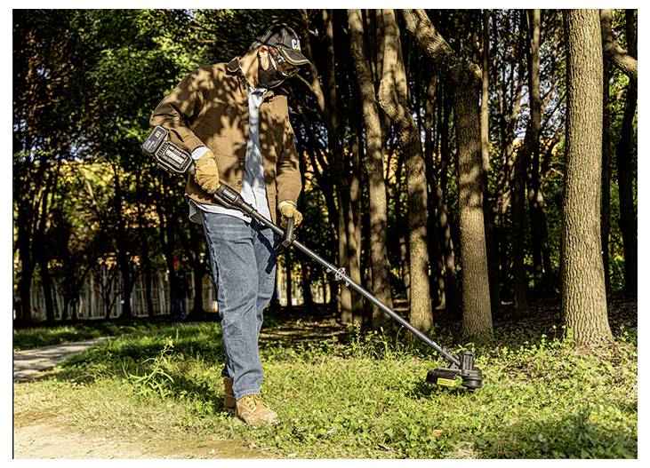 Person using a string trimmer to cut grass along a wooded path in a park.