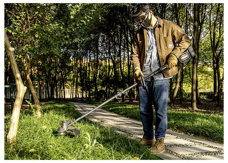 Man using a cordless string trimmer to cut grass along a park path surrounded by trees.