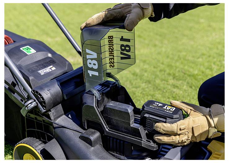 Gloved hands insert an 18V battery into a lawn mower power slot outdoors on grass.<br>