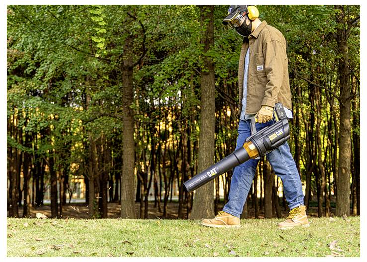 Person in protective gear using a battery-powered leaf blower in a grassy wooded area.