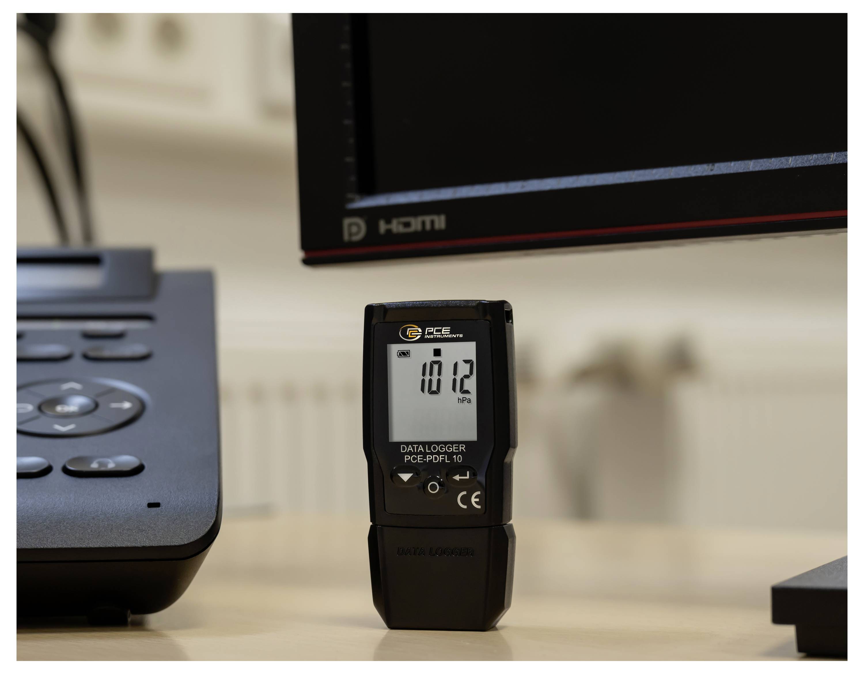 A handheld digital data logger stands on a desk in front of a computer monitor, showing a reading of 18.2.