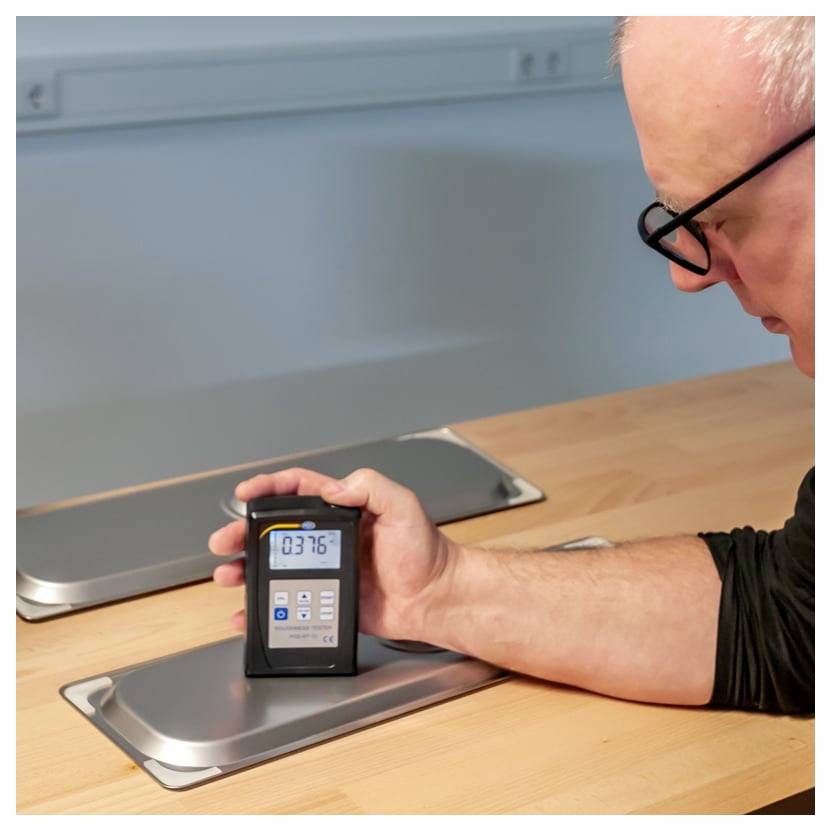 Man holding a handheld digital meter over a metal panel on a wooden table, apparently measuring a surface reading.