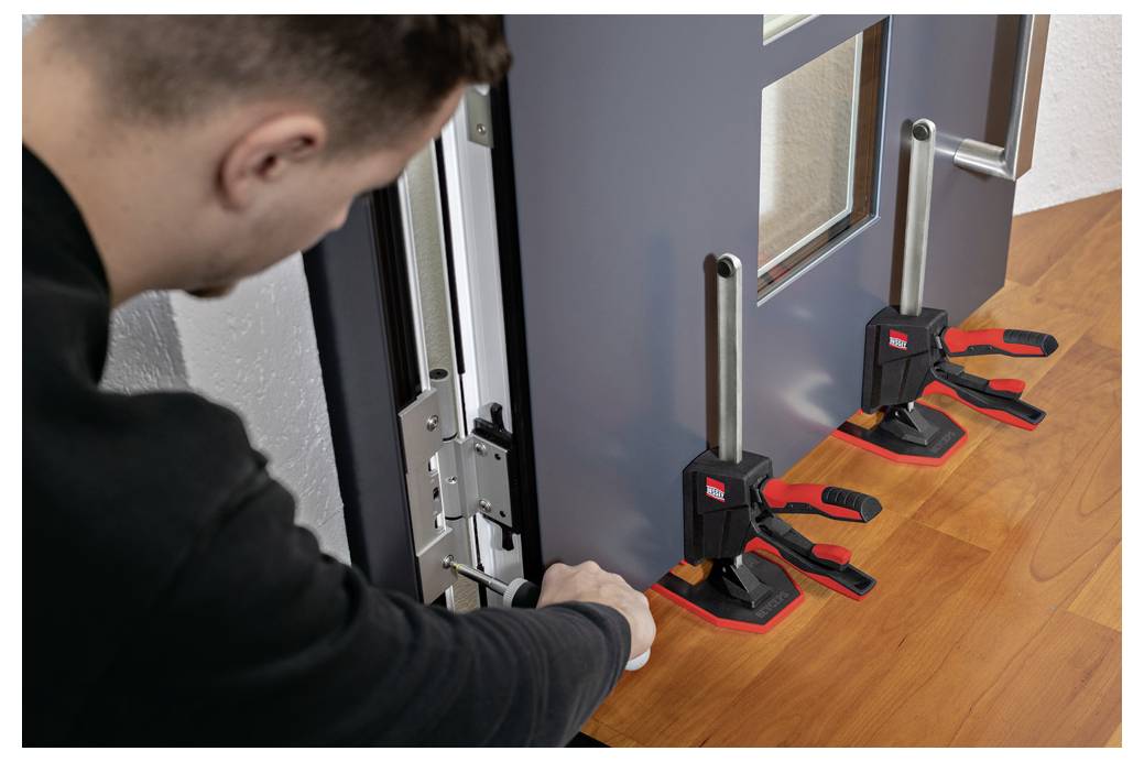 Person adjusting and clamping a gray door or panel to a wooden floor with three black-and-red bar clamps.