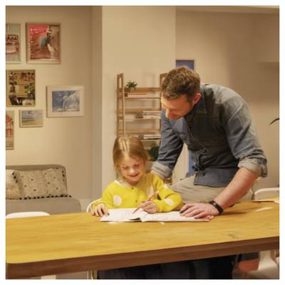 Man helps a young girl write at a table in a home, both focused on the paper.