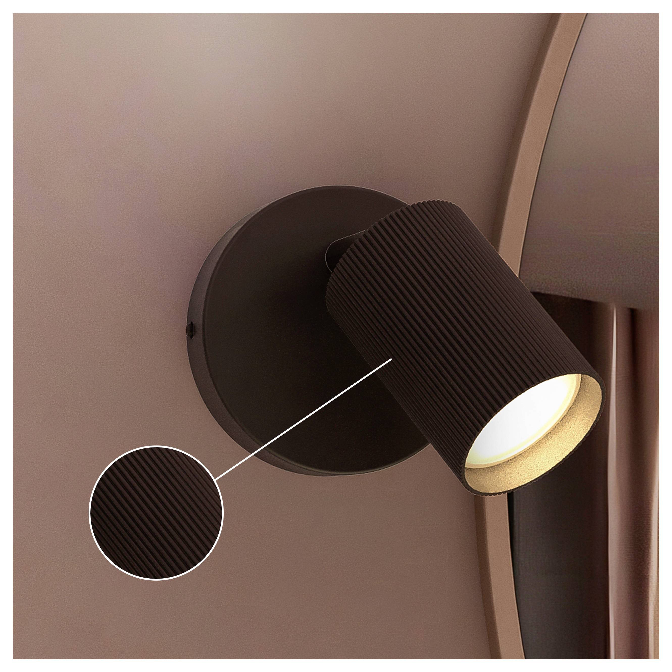 Black cylindrical wall spotlight with ribbed shade, lit and angled downward on a brown wall.
