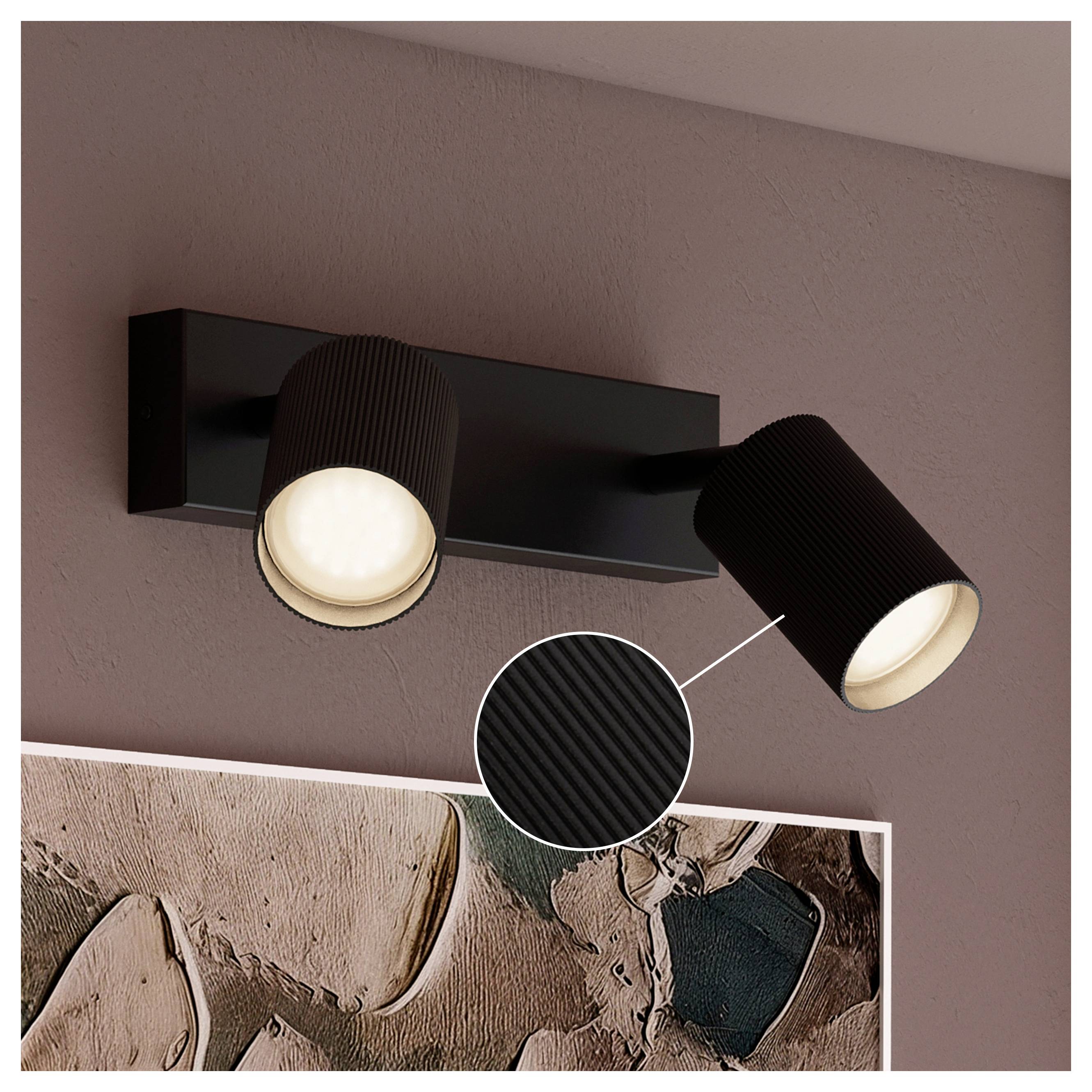 Black two-light wall sconce with adjustable spotlight heads illuminating a framed abstract artwork on a mauve wall; inset shows ribbed surface detail.