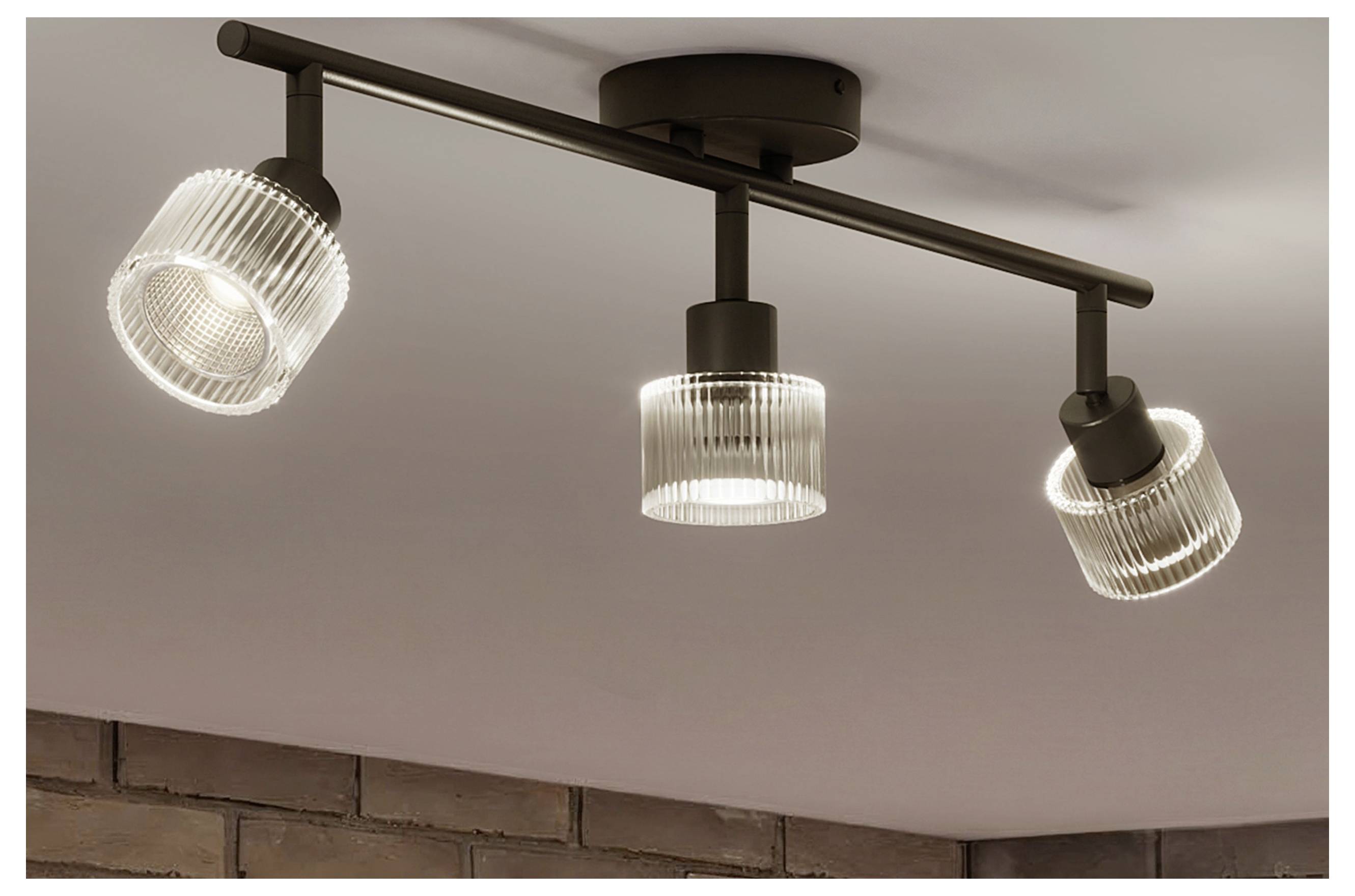 Three-light black ceiling track fixture with adjustable ribbed glass shades mounted above a brick wall.