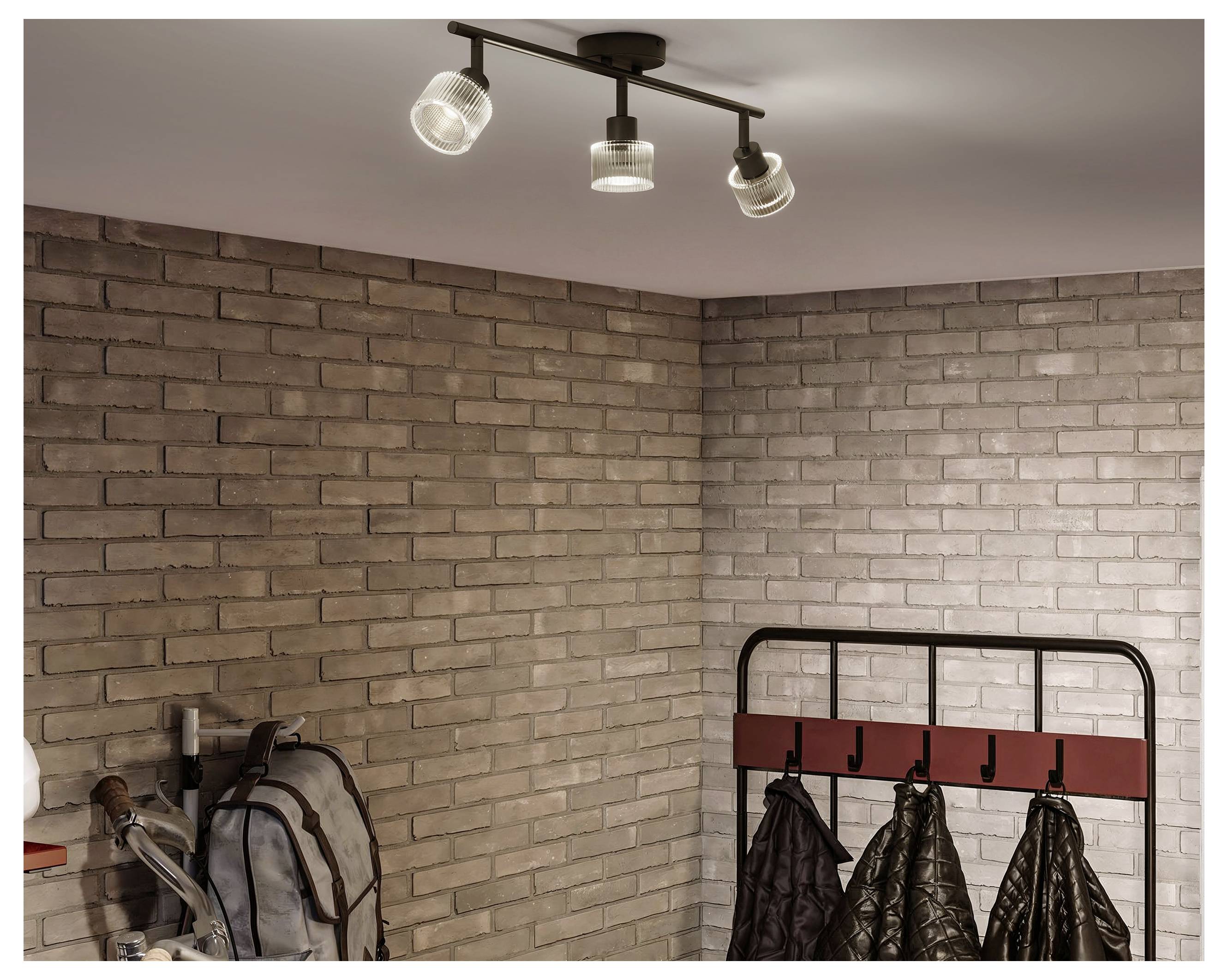 Empty corner with beige brick walls, ceiling spotlight, backpack, and coat rack holding jackets.