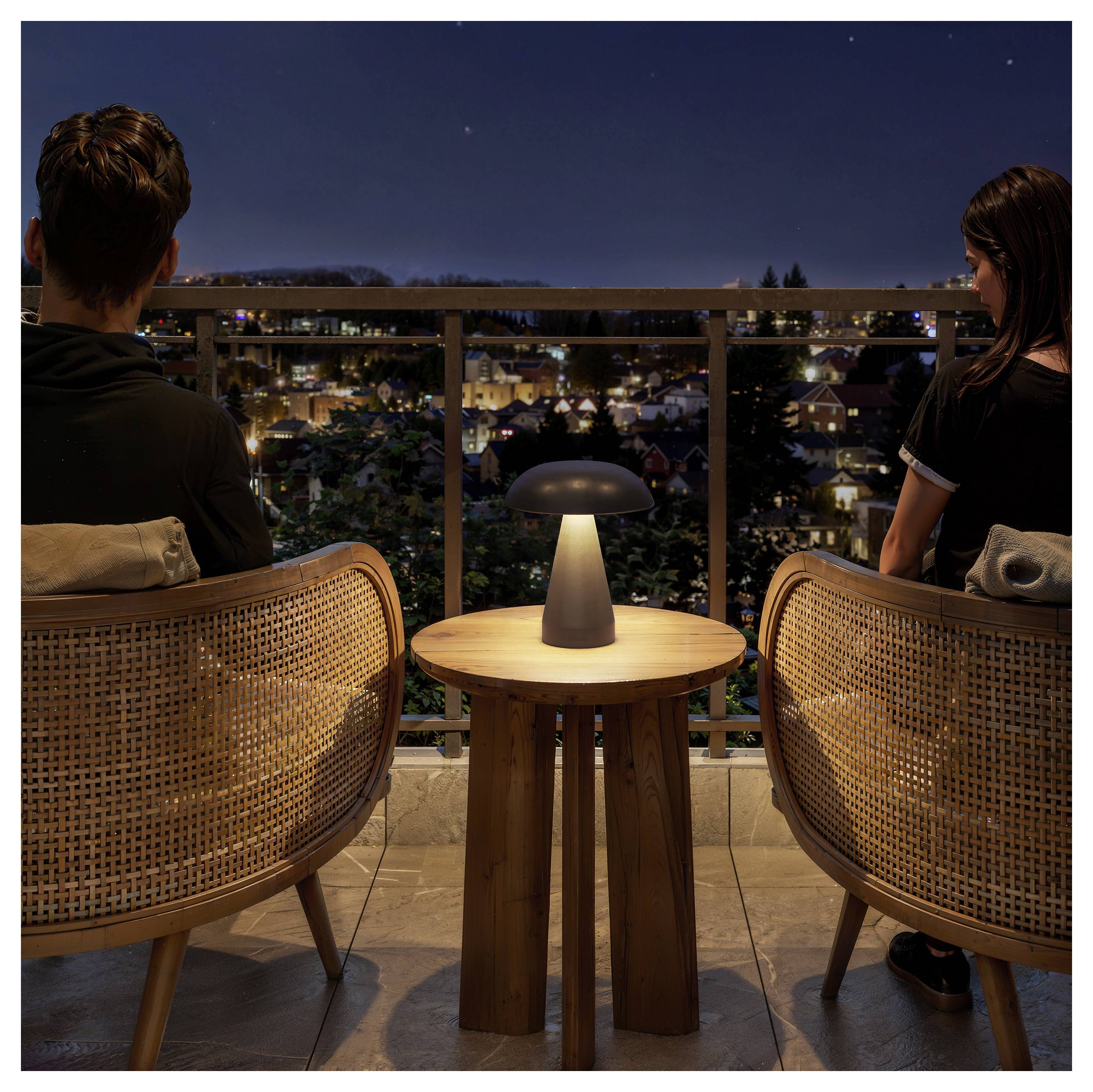 Two people sit on a balcony at night, facing a lit city neighborhood with a small lamp on the table between them.<br>