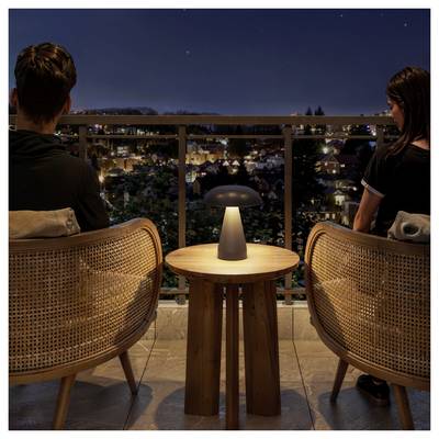 Two people sit on a balcony at night, facing a lit city neighborhood with a small lamp on the table between them.<br>