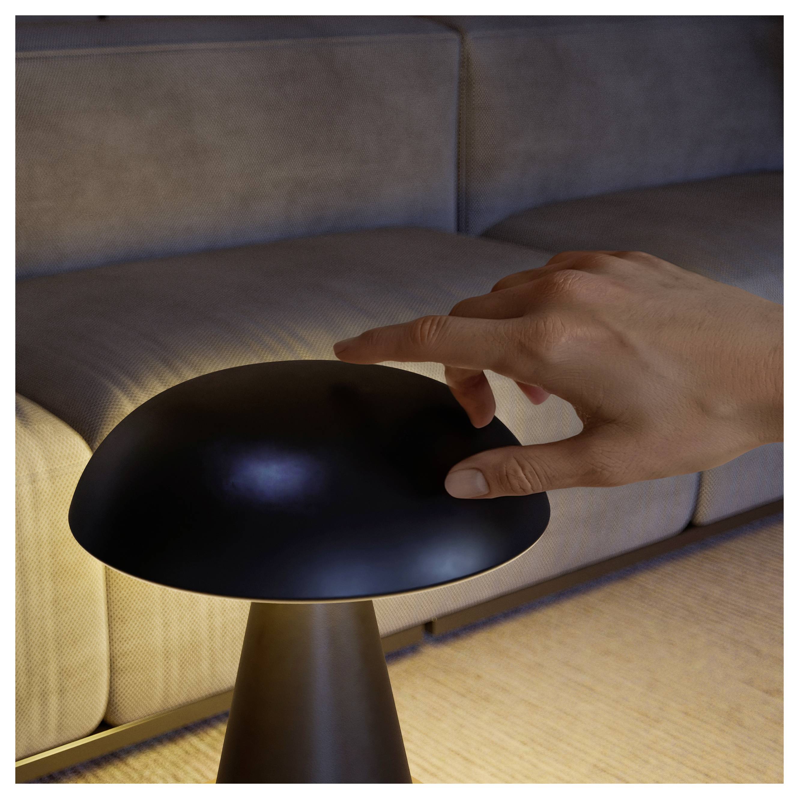 Hand hovering over a dome-shaped black lamp on a table beside a sofa in a softly lit room.