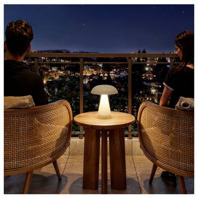 Two people sit on a balcony at night, facing a lit cityscape, with a small glowing table lamp between their chairs.