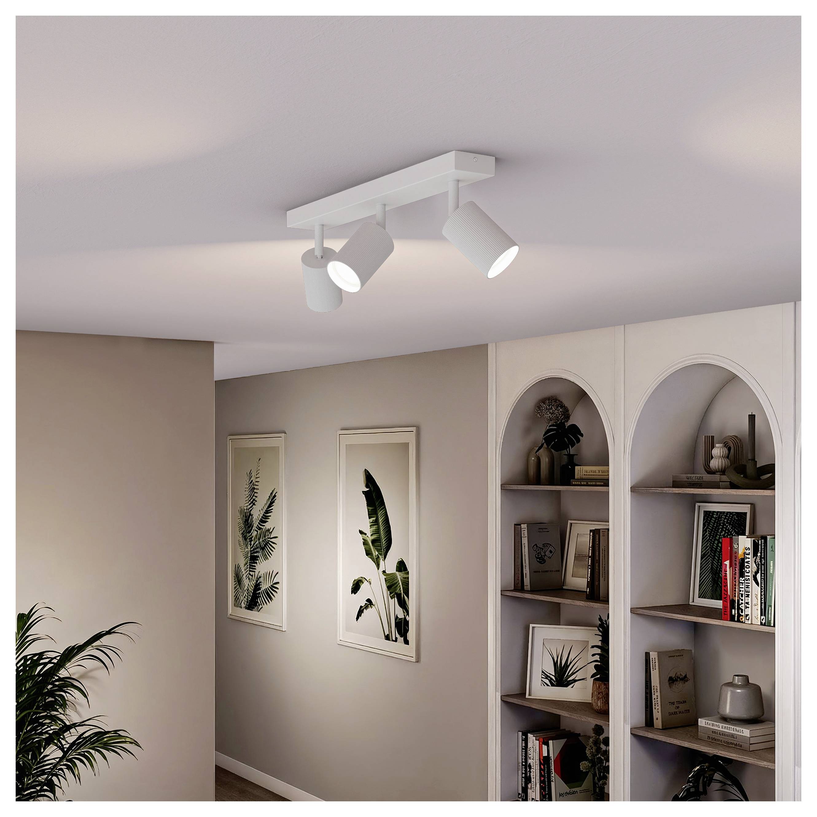 White ceiling track light with three adjustable spotlights in a beige room, beside arched wall shelves and framed plant prints.<br>