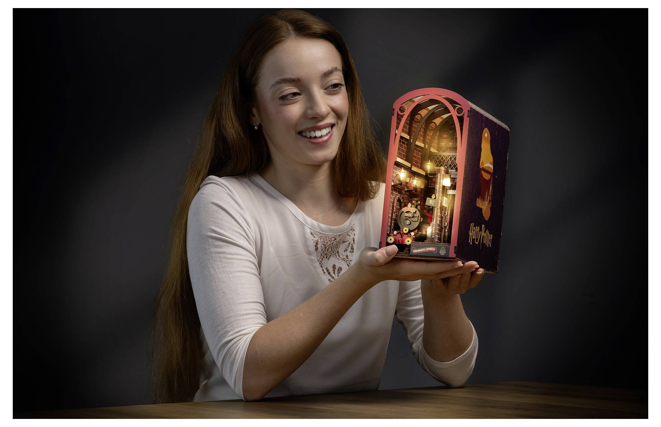 Smiling woman presents an illuminated miniature building model with a lit arched doorway on a table against a dark background.