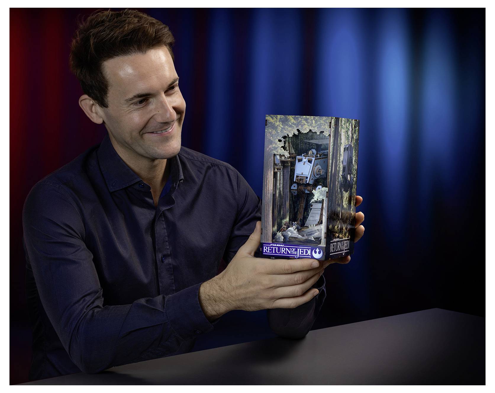 Man smiles while holding up a 'Return of the Jedi' collectible box at a table against a dark red and blue backdrop.