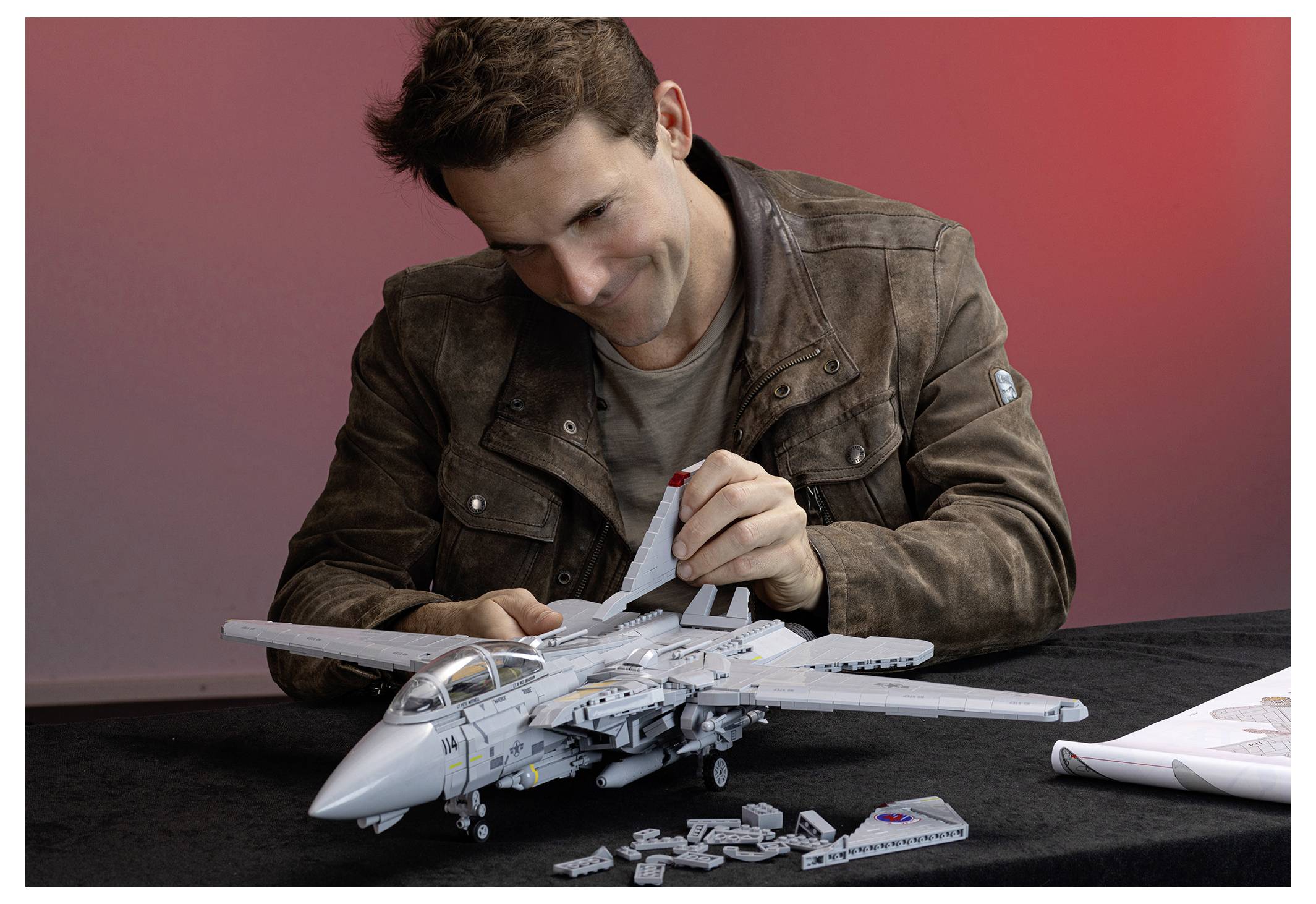 Man assembling a detailed model fighter jet on a table, with small parts scattered around and a red gradient background.