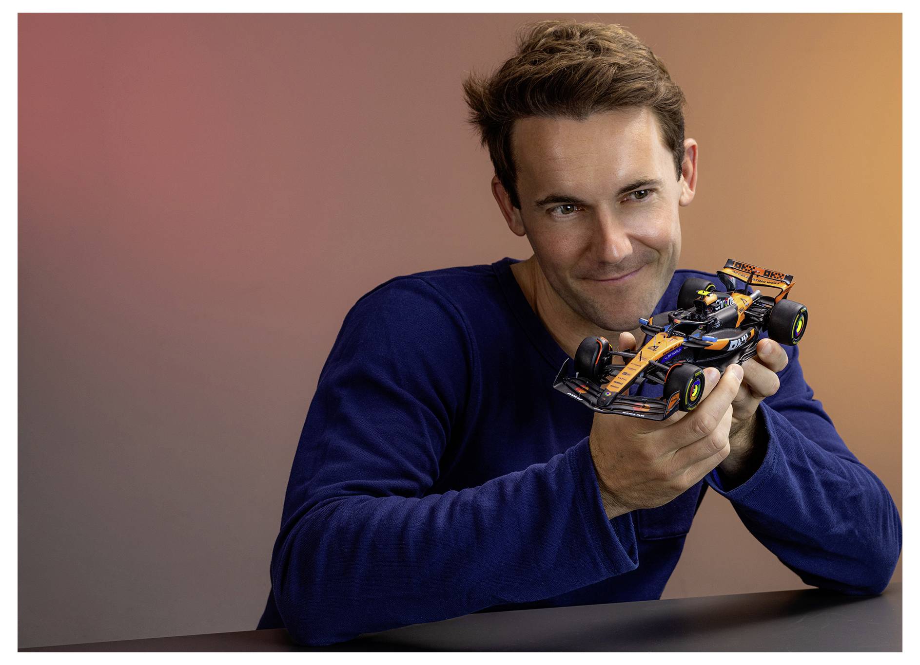 Man holds a detailed orange-and-blue Formula 1 race car model, smiling against a plain studio background.<br>