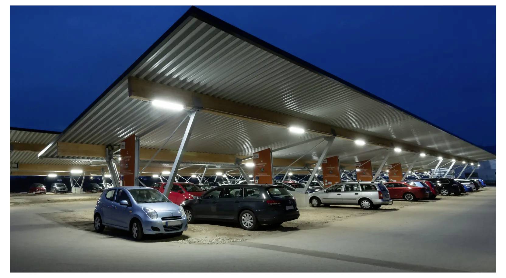 Nighttime photo of a modern solar-panel carport at a parking lot, with several cars parked beneath the illuminated canopy.