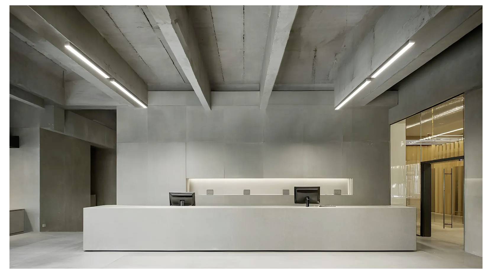 Minimalist modern office reception desk in a concrete interior with two computer monitors and recessed lighting, viewed from the lobby.
