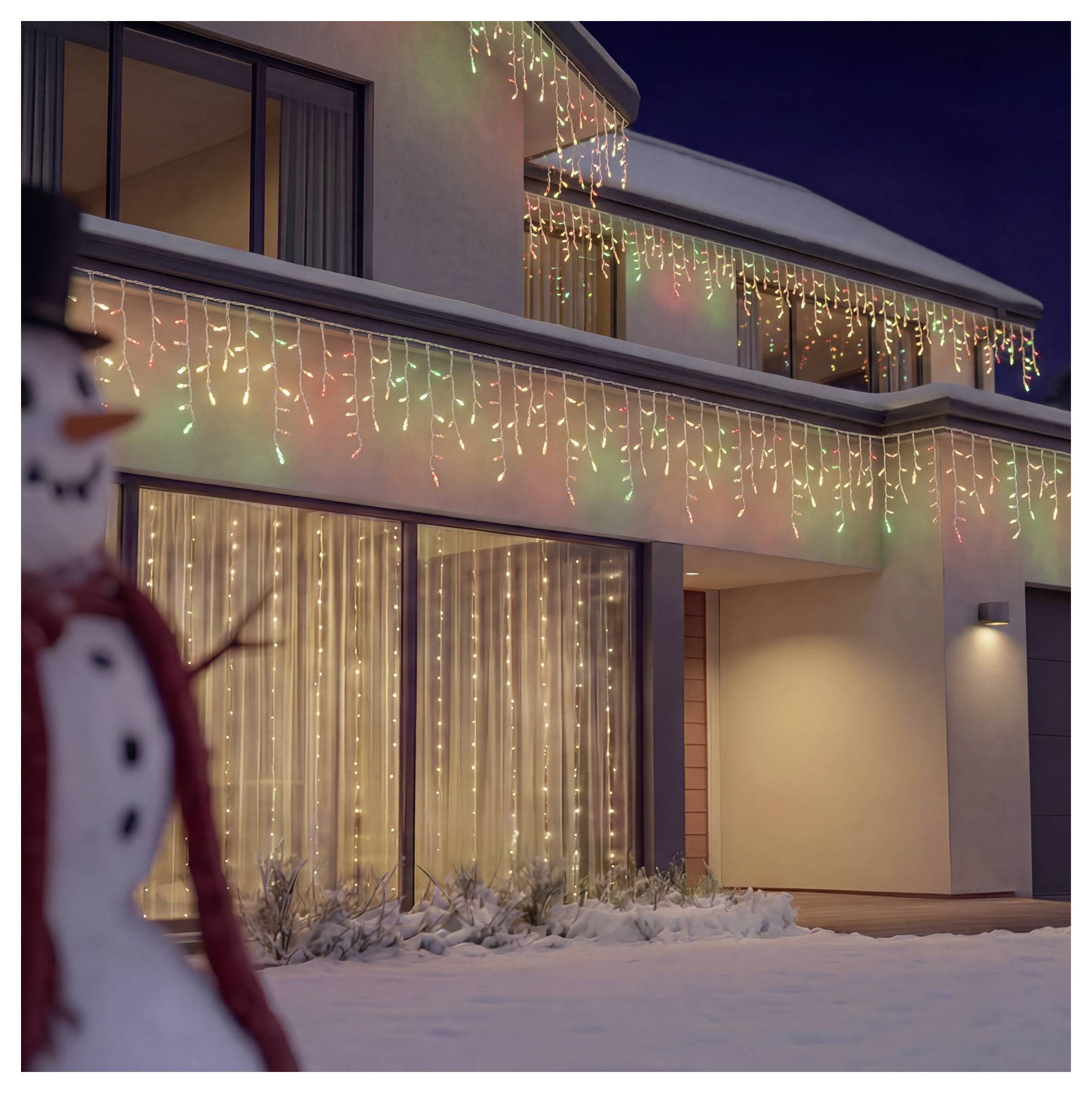 Snowy modern house at night decorated with multicolored holiday string lights, with a blurred snowman in the foreground.