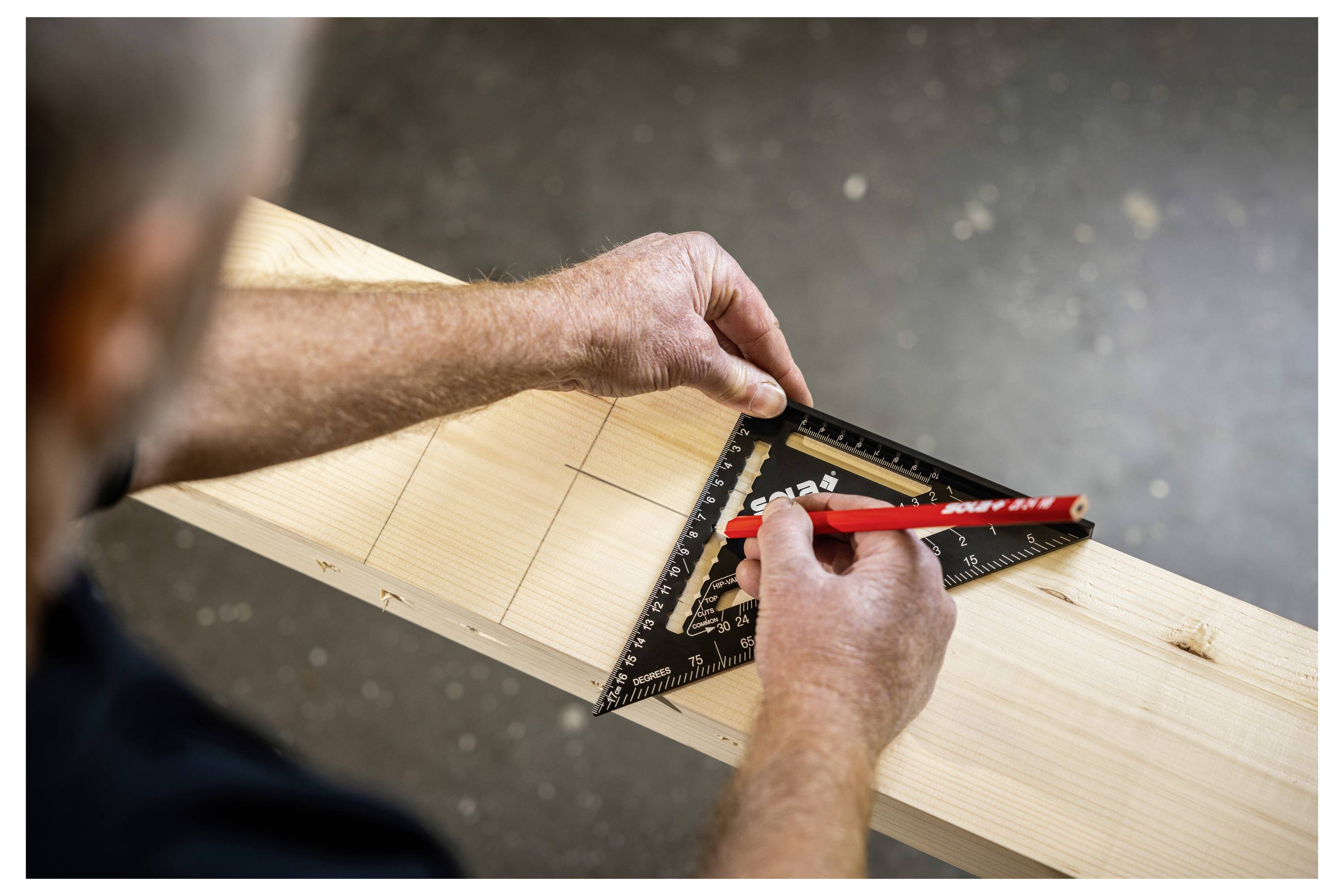 Hands marking a cut line on a wooden beam with a carpenter’s square and red pencil.<br>