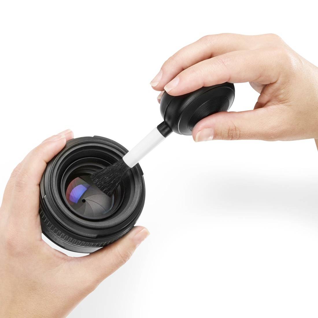 Hands using a blower brush to clean dust from a camera lens on a white background.