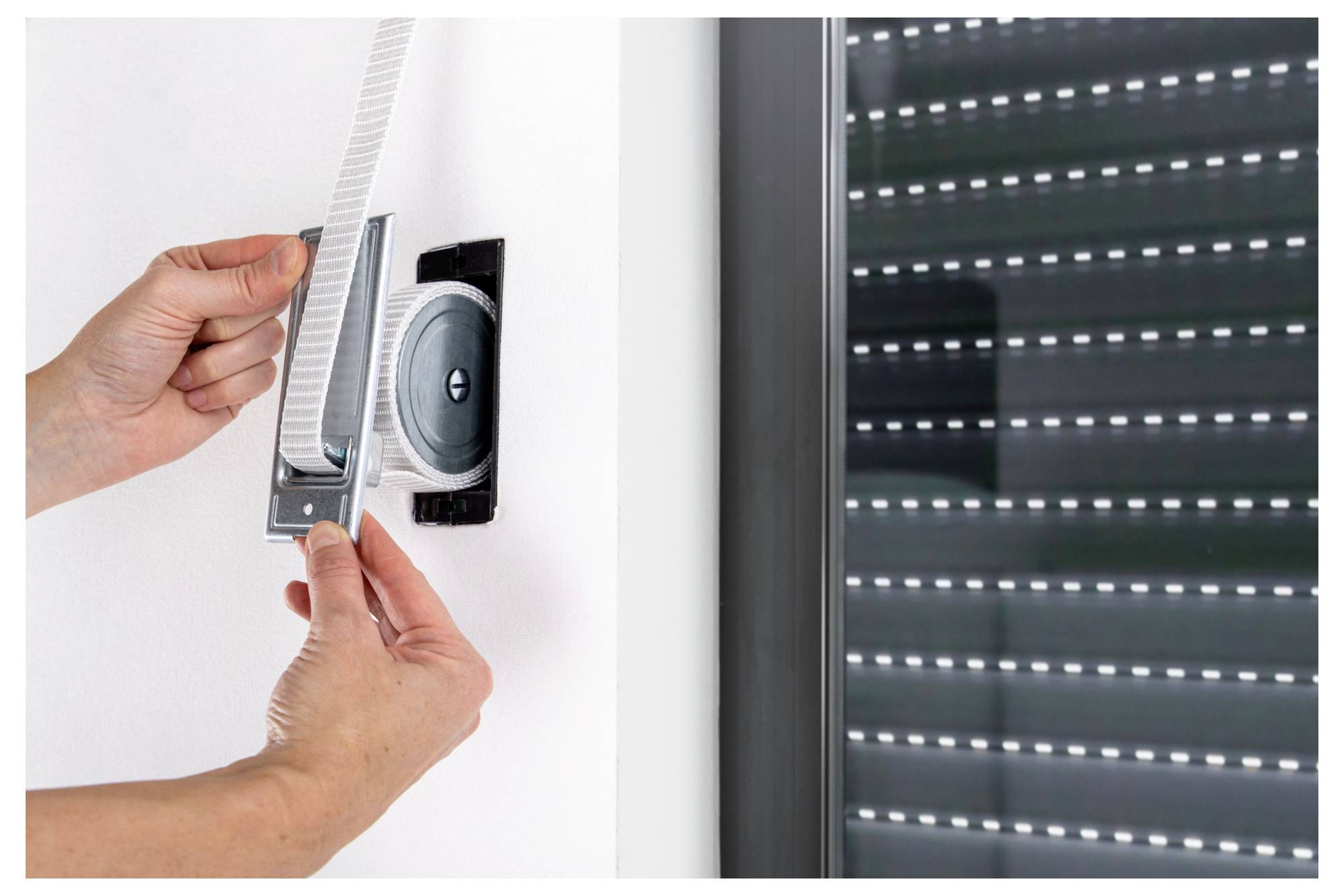 Hands installing a white roller blind cassette onto a window above a dark shuttered window frame.